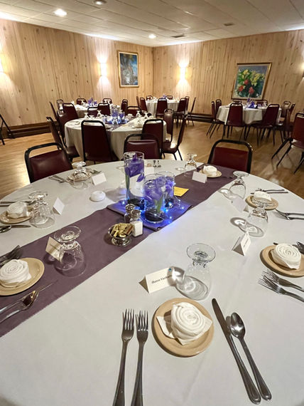 Round banquet tables set with white linens, purple runners, and glass centerpieces in Holiday Acres Resort event room.