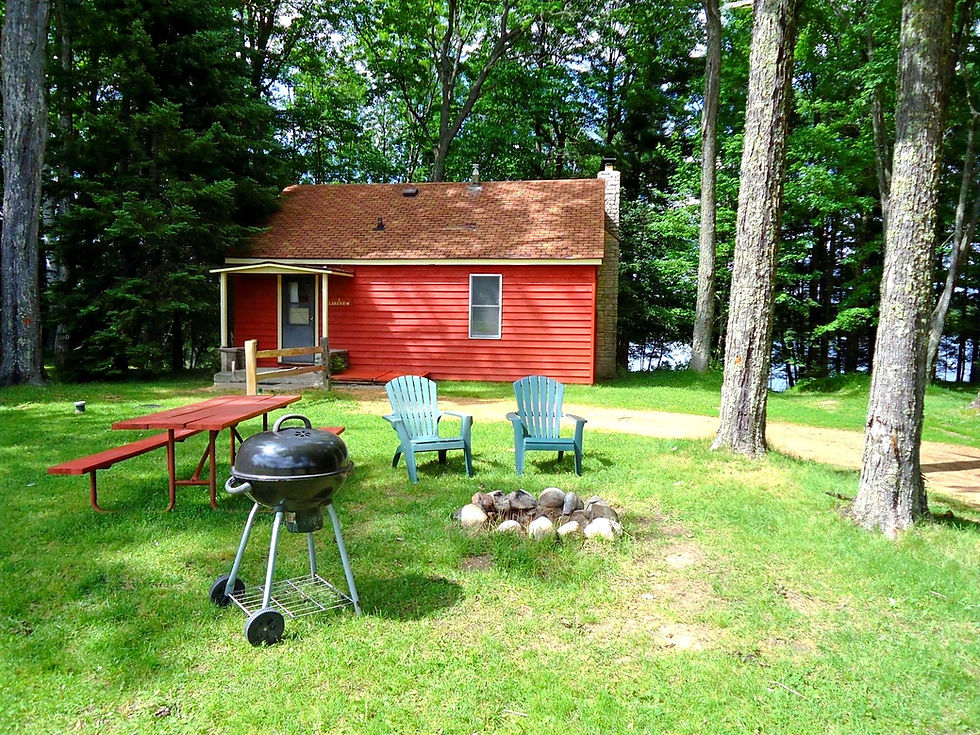 A red cabin with a sloped roof surrounded by grass and trees, with a picnic table, fire pit, and Adirondack chairs in the foreground.