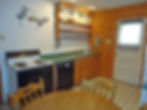 A kitchen with wood and white walls, a white stove, dishwasher, coffee maker, and open shelves stocked with dishes. A wooden dining table is partially visible.