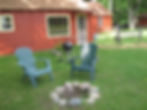 A grassy outdoor area with two green Adirondack chairs, a circle of firepit stones, and a charcoal grill in front of Meadowview Cottage.