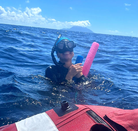 Child in snorkel gear holds pink float near boat, distant island.
