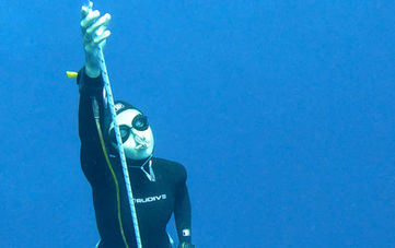 Person in black wetsuit freediving, holding descent rope in deep blue ocean.