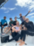 Group of seven people in wetsuits smiling on a boat under blue sky.