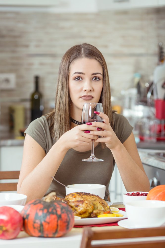 A woman sits alone with a glass of wine and chicken dinner, looking sad. Some are alone at Christmas, amidst the hype of a happy family season. Loneliness is a holiday stress.