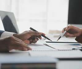 Two people across the table from one another gesture to papers with pens, conversing. 