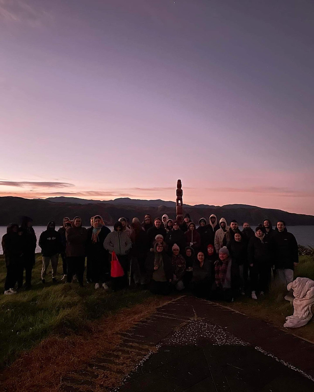 Group of rangatahi dressed warmly stand together with a pou that stands at Oruaiti. Behind them is the harbour, hills and a beautiful early morning sky.
