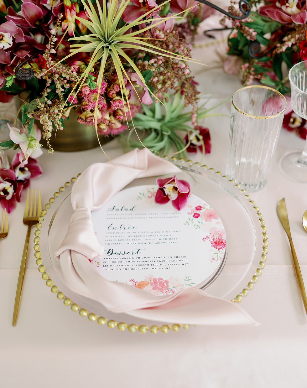 Table setting with magenta and green orals and beaded gold rimmed charger with circular designed menu and elegant knotted silk napkin