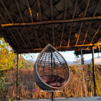 Cozy hanging egg chair under a thatched bamboo roof at Noel’s Camping Roing with greenery and hills