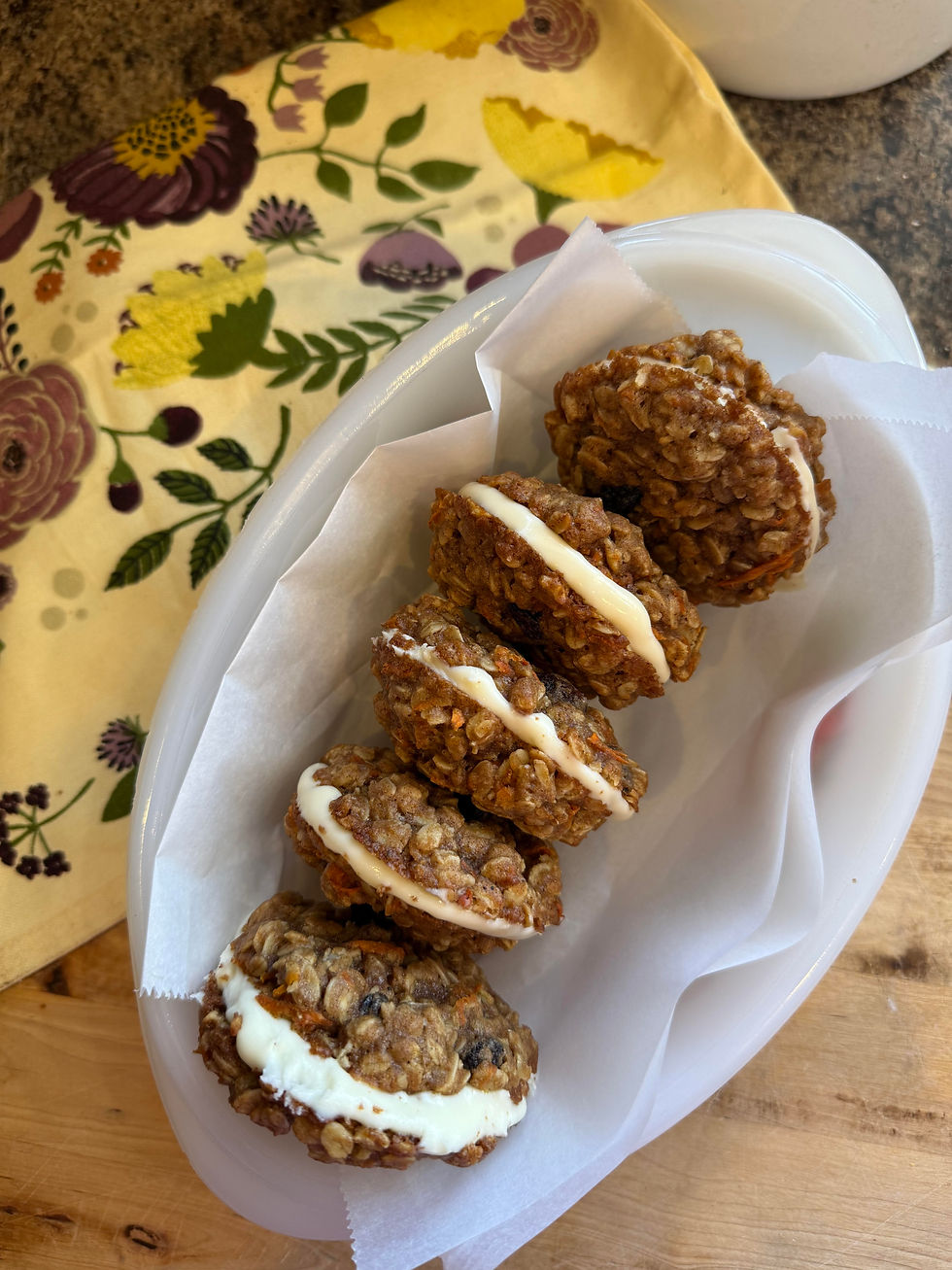 Oatmeal cream pies with frosting filling on wax paper in a container. Floral-patterned cloth in the background. Cozy and inviting.