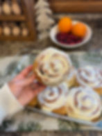 Hand holding a frosted cinnamon roll above a tray of more rolls. Background has a bowl of oranges and cranberries, and a holiday theme.