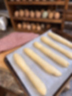 Four unbaked baguettes on parchment-lined tray, kitchen counter; egg rack with various eggs in background, red checkered cloth nearby.