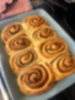 Freshly baked cinnamon rolls in a blue dish, golden brown with swirls. A red checkered cloth is in the background, suggesting a cozy kitchen.