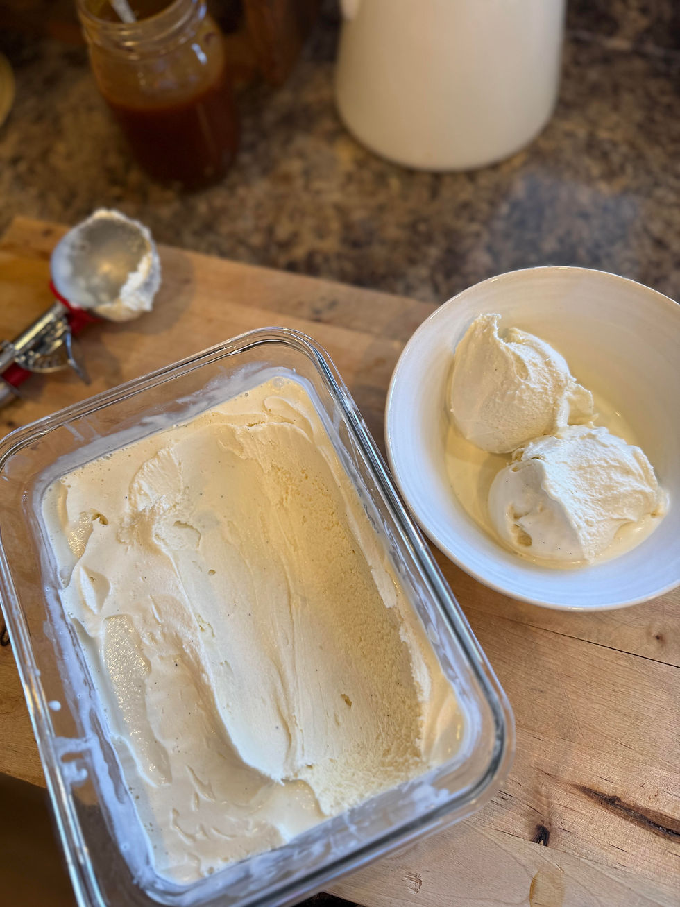 Ice cream in a glass container and scooped into a white bowl on a wooden counter. Metal scooper and jar in the background. Creamy texture.