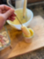 Hand dipping a sausage on a stick into a jar of batter. White bowl and utensils on a wooden surface in the kitchen setting.