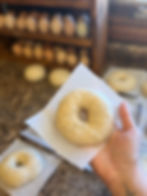 A hand holding an uncooked bagel on parchment paper, with more bagels and a wooden egg rack in the blurred background. Warm, cozy mood.