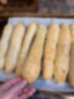 Hand holding a golden-brown garlic breadstick on a baking tray with parchment paper. Six other breadsticks are lined up. Kitchen counter in background.