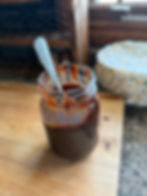 A jar of dark sauce with a decorative spoon on a wooden counter, lid off. Background includes a window and a fabric-covered item.