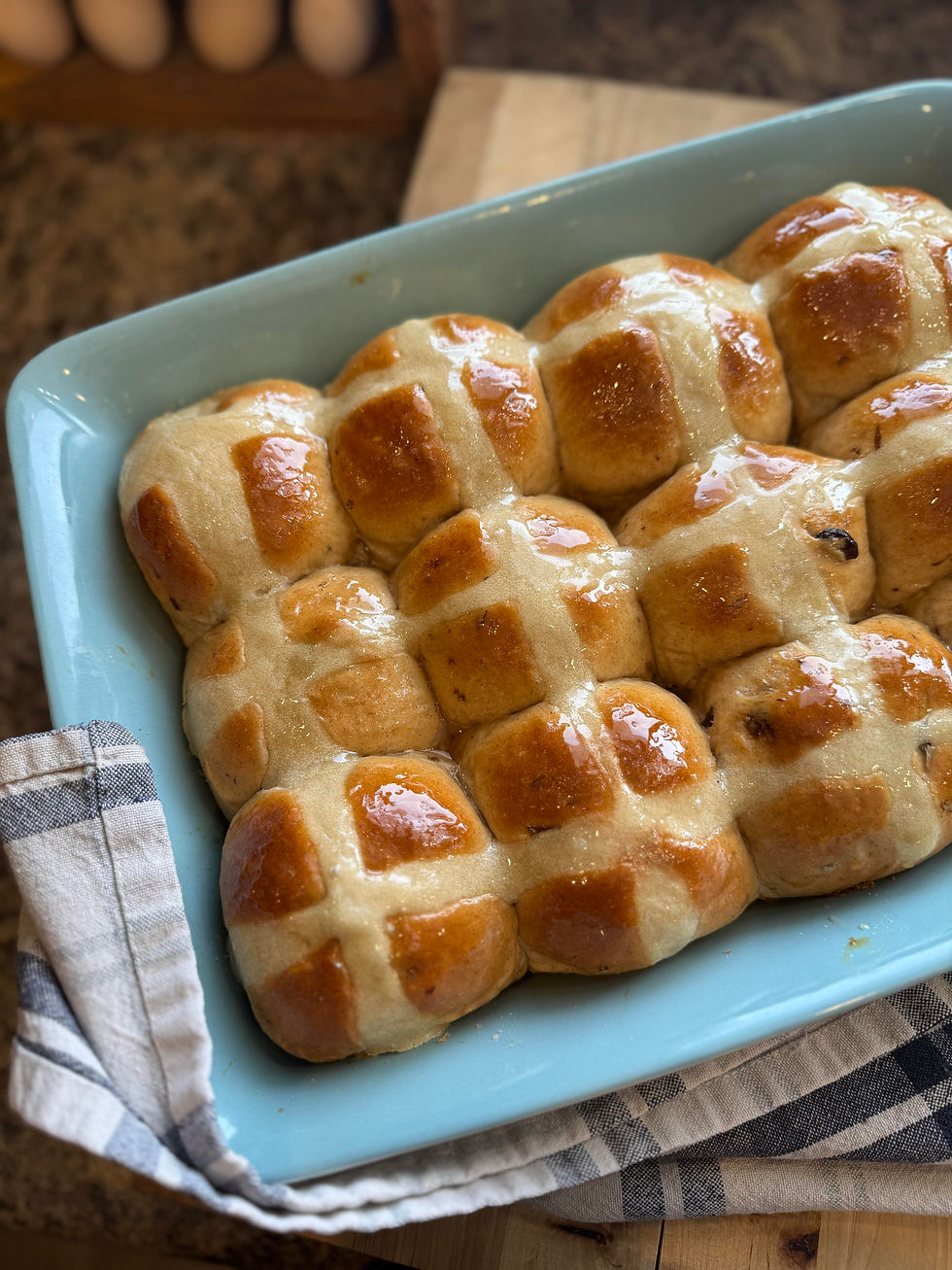 Golden brown hot cross buns with icing in a light blue baking dish on a checkered cloth. Eggs are visible in the background.