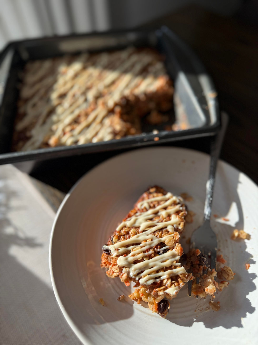 Granola bar drizzled with white icing on a white plate with a fork, sitting in front of a baking tray on a sunlit table.