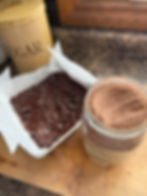 Chocolate batter with chips in a parchment-lined pan next to a jar of cocoa powder. Sugar canister in the background on a wooden counter.