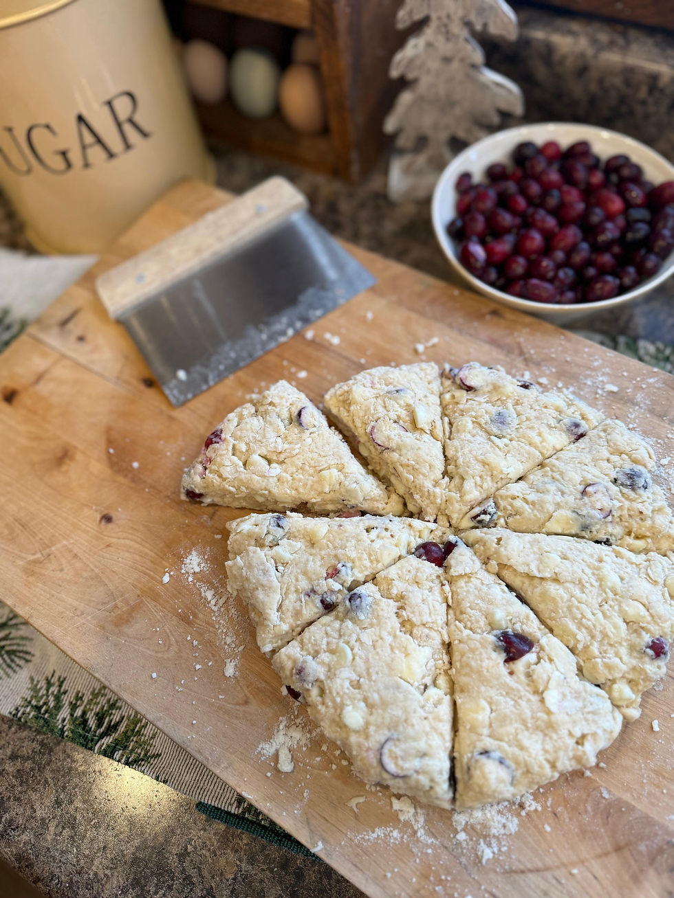 Scones with cranberries on a floured wooden board, metal scraper nearby. Background: sugar container, bowl of cranberries, eggs, pine tree decor.