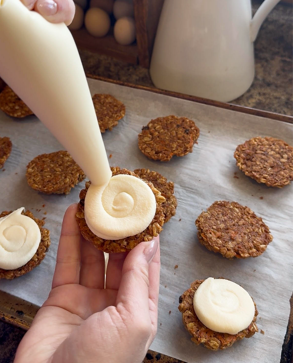 Piping cream onto oatmeal cookies on a parchment-lined tray, with some sandwiched by cream. Warm-toned cookies with visible carrot bits.