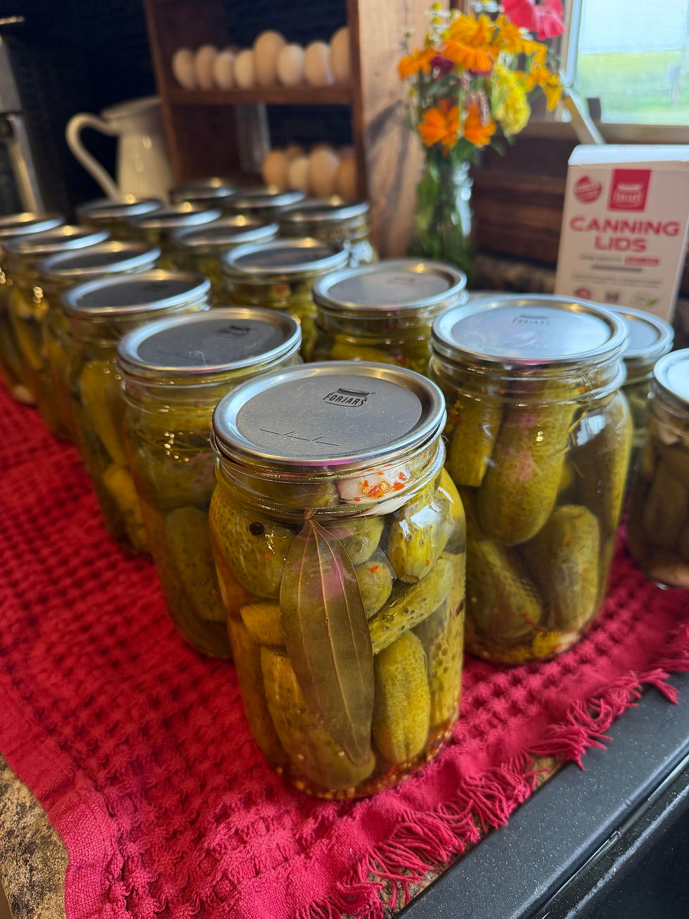 Jars of homemade pickles and spices line a red cloth in a kitchen, beside a box labeled Canning Lids and flowers.