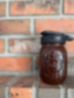 A glass mason jar filled with homemade chocolate syrup sits on a brick ledge. The jar has a black lid and embossed text on the front. Background: red bricks.