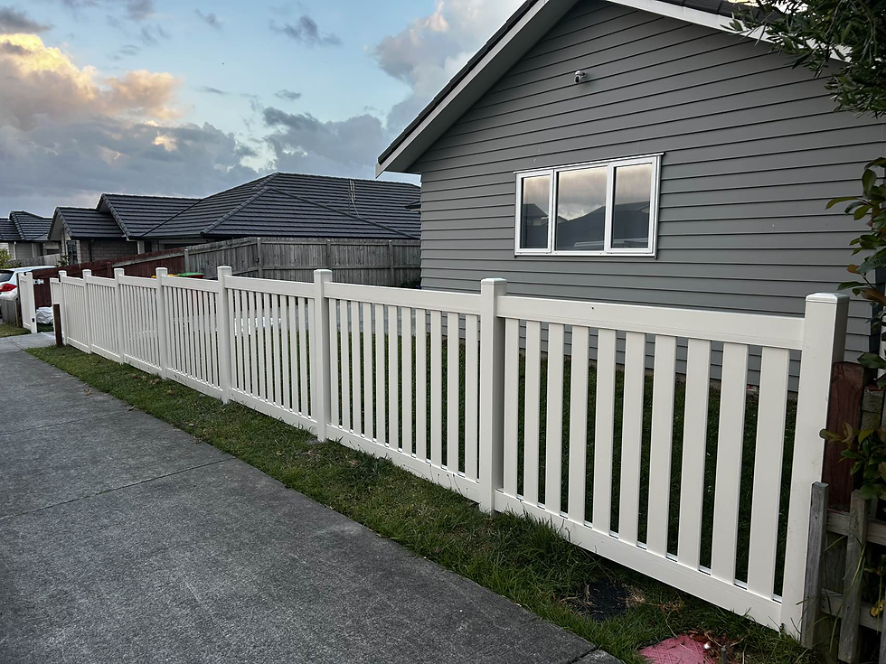 White picket fence lines a suburban street, fronting a gray house under a cloudy sky with evening light. Calm, peaceful neighborhood scene.