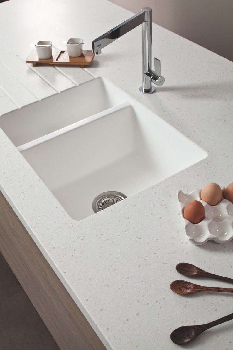 White kitchen counter with a sleek chrome faucet over a double sink. Nearby are a wooden tray with cups and an egg holder with brown eggs.