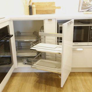Modern kitchen corner with open white cabinets, revealing pull-out trays and a microwave. Wooden floor, countertop with cutting board.