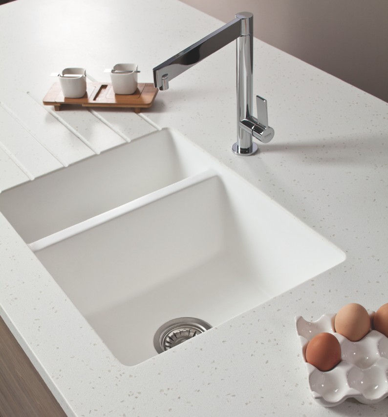 White kitchen counter with a sleek chrome faucet over a double sink. Nearby are a wooden tray with cups and an egg holder with brown eggs.