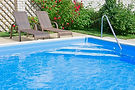 Two brown wicker lounge chairs sit on a patio beside a clear blue swimming pool, with green shrubs and blooming red flowers in the background.