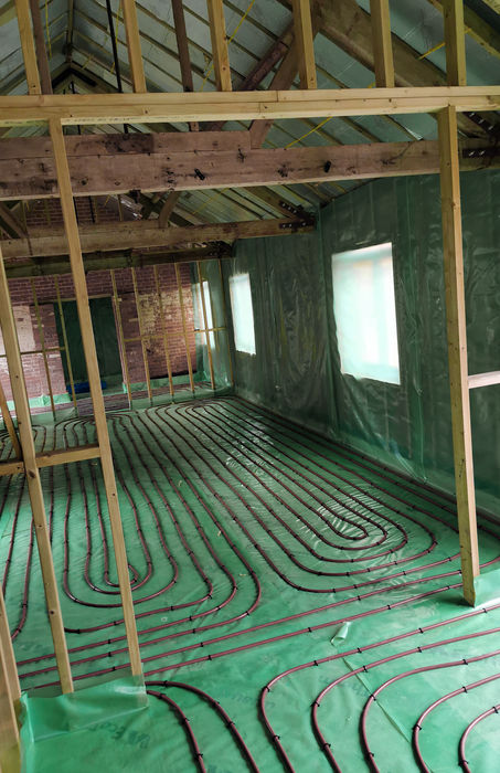 Exposed wooden framing inside a house under construction, with red underfloor heating pipes laid out in loops over a green insulating layer. Walls are covered in green plastic sheeting, and windows are covered.