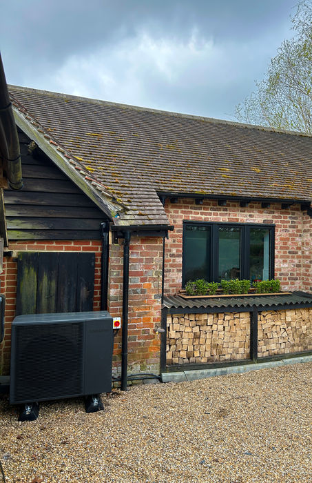 A brick building with a shingled roof, black-framed window, stacked firewood under the window, and an outdoor heat pump unit on a gravel surface.
