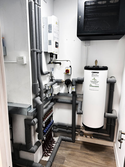 A utility room with a water heater, insulated pipes, control panels, valves, and gauges mounted on white walls; a black cabinet is fixed high on the right wall above the water heater.