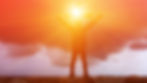 a man on a hilltop with arms raised toward a setting sun and the image of the cross.