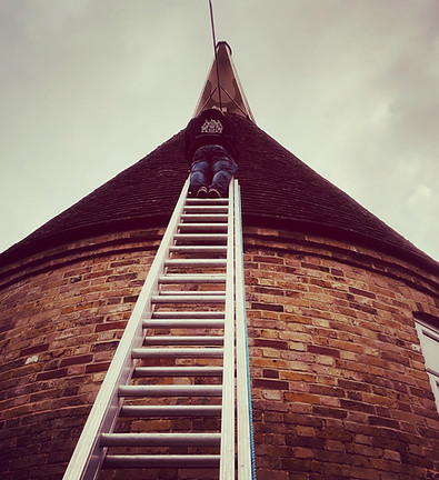A specialist roofer from G&S Oast Cowl Refurbishment climbing a ladder to repair a traditional brick oast house roundel in Kent.