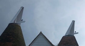 Two pristine white traditional oast cowls with black horse finials on top of Kent peg tile roundels against a cloudy sky in Kent.