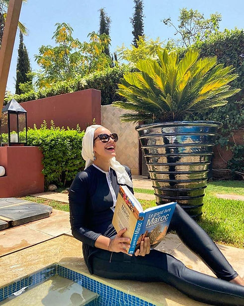 photo d'une femme en burkini hijab noir, elle rit aux éclats en lisant un livre sur le Maroc