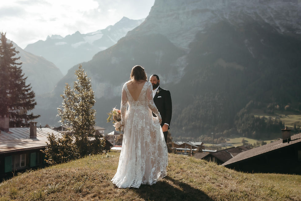 Bride in white gown and groom in black tux stand on a grassy hillside with alpine chalets in the background in Grindelwald