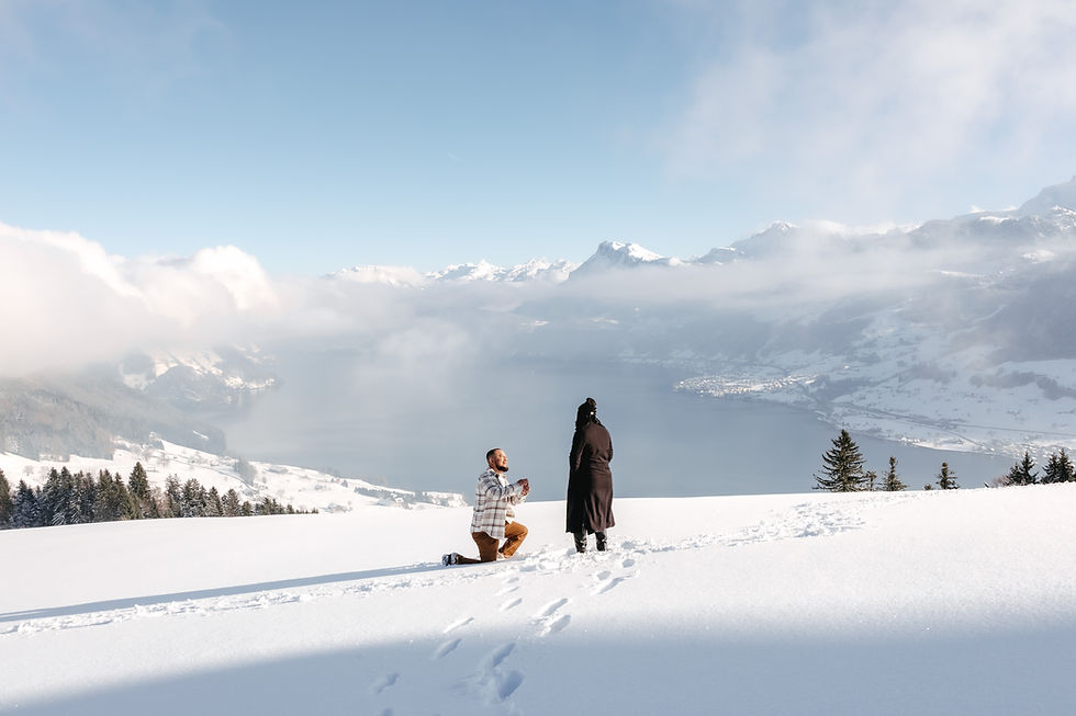 Man kneels in snow proposing to a bundled-up woman on a mountain overlook above a misty lake and snowy peaks. Villa Honegg's meadow covered in snow during a proposal.