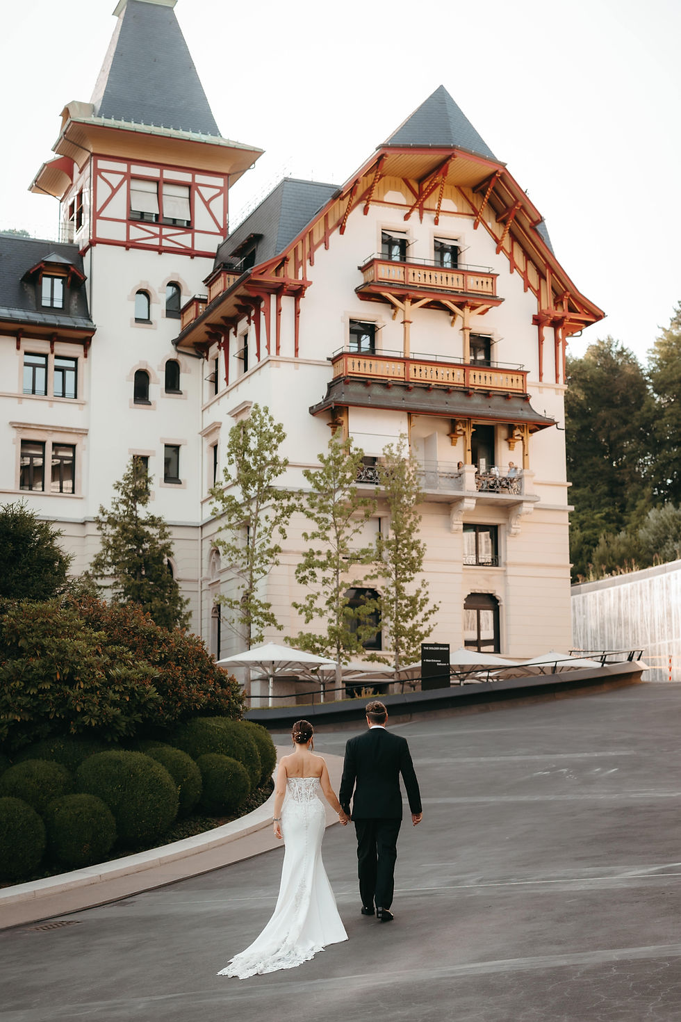 Bride in white gown and groom in black suit walk hand in hand toward a grand castle-like hotel with red trim. The Dolder Grand hotel exterior, Zurich, above the city, wedding venue Switzerland.