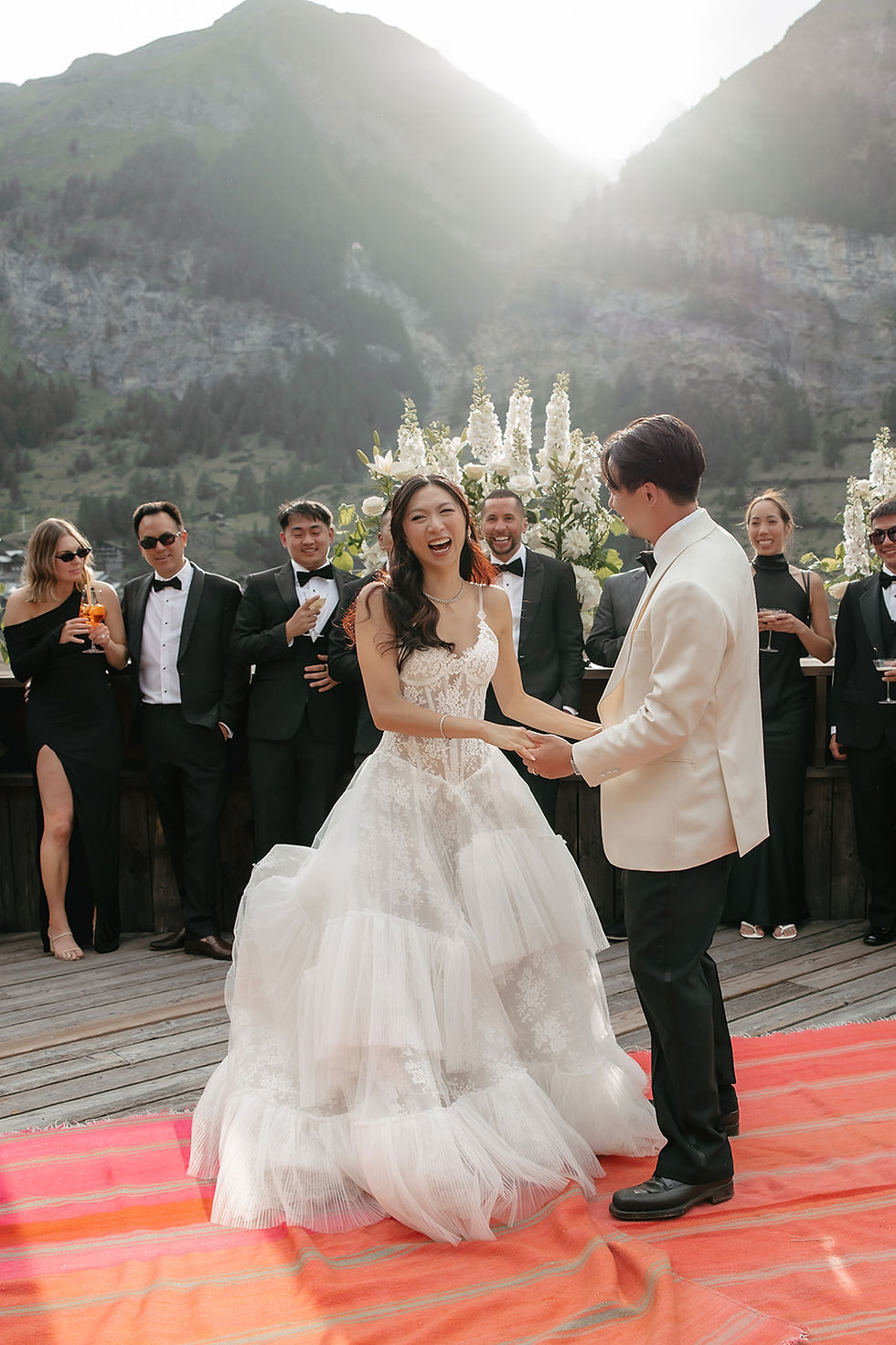 Bride in lace wedding dress smiles while dancing with groom in cream jacket outdoors, with guests and white flowers behind them. Couple's first dance at their destination wedding at Cervo Zermatt