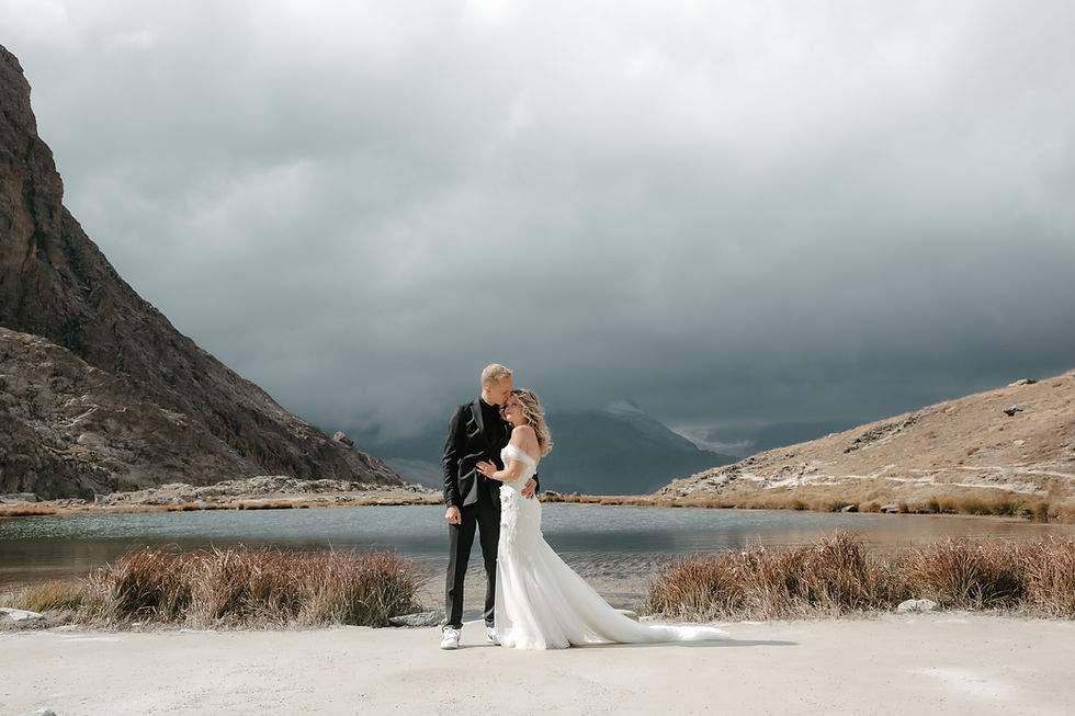 Bride in white gown and groom in black suit embrace by a mountain lake under dark cloudy skies, romantic and dramatic mood. Couple on their wedding photoshoot in Zermatt, in front of the Matterhorn at the Gornergrat.