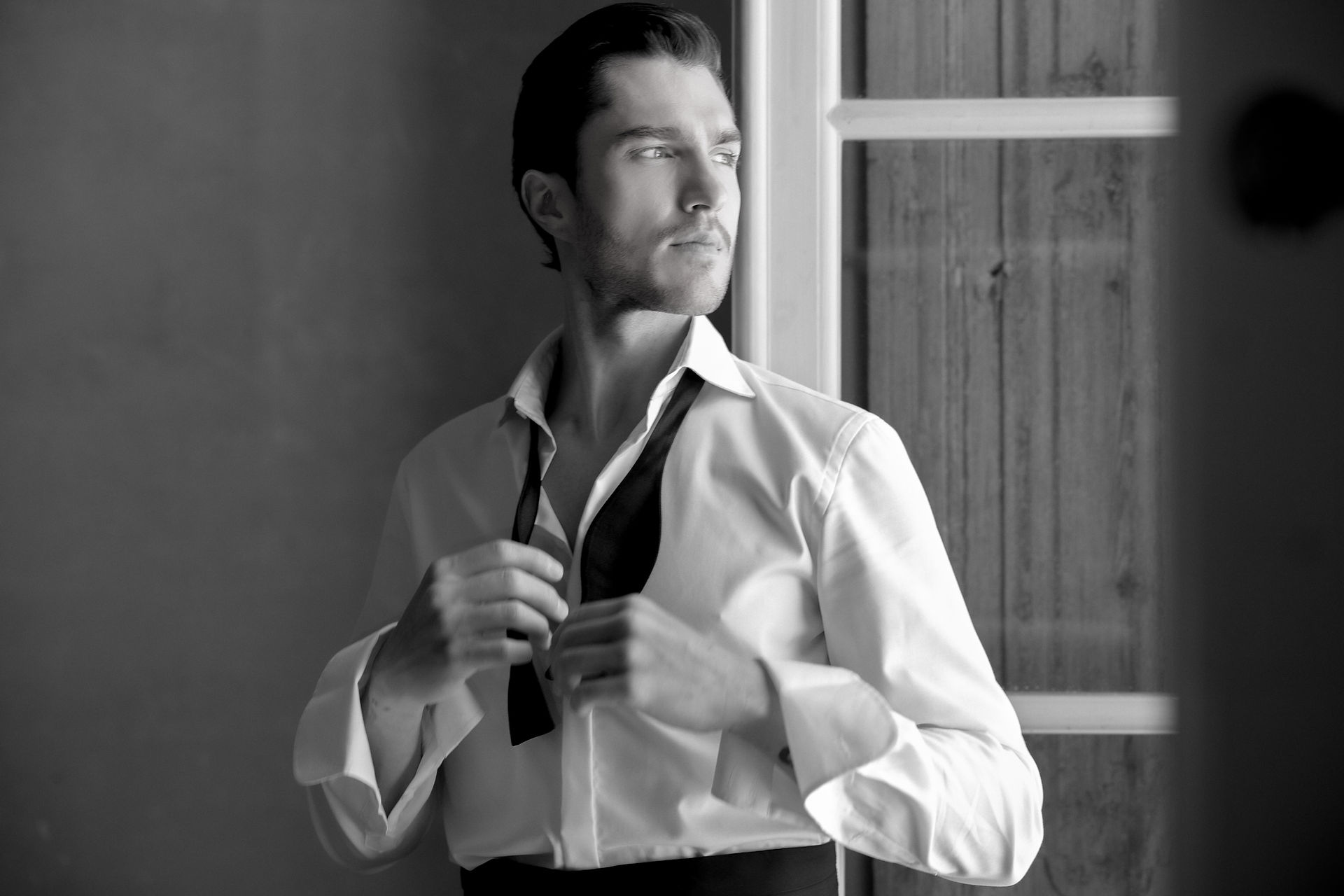Groom elegantly posing during getting ready. Groom buttoning his shirt. Black and white. 