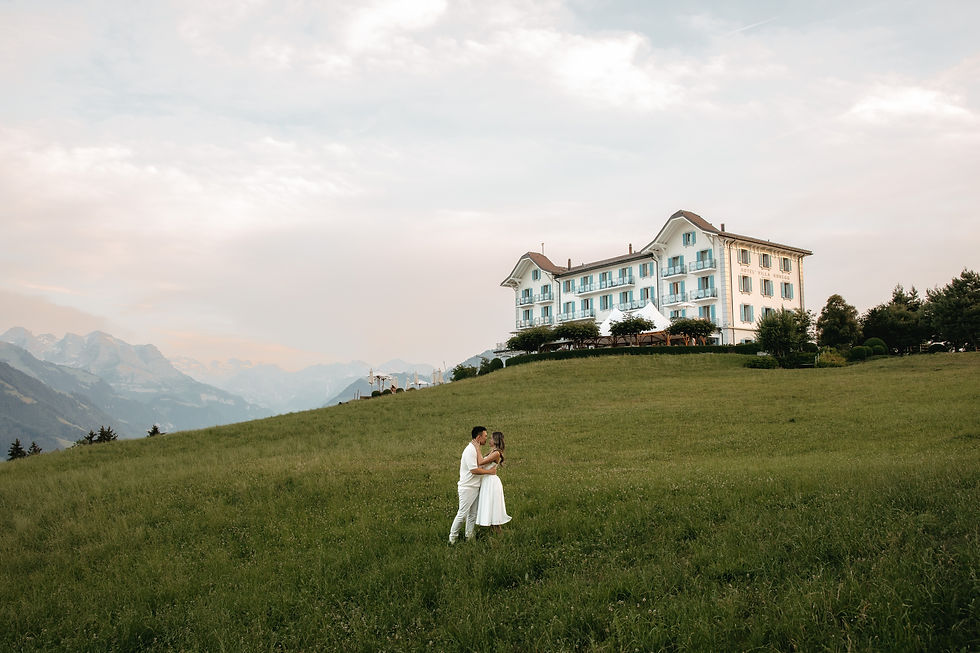 Couple in white embrace on a grassy hill by Hotel Villa Honegg, with alpine mountains and cloudy sky in the background.