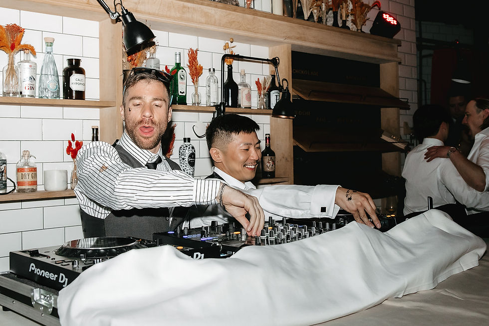Two men in dress shirts DJing at a Pioneer DJ booth in a lively bar, with bottles on shelves and partygoers in the background. Groom's friends as djs during a party at Sala Ferdinand at Cervo in Zermatt