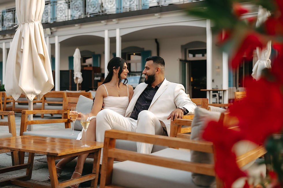 Couple in white outfits sit at an outdoor patio table, smiling at each other with drinks, red flowers blurred in foreground, after their marriage proposal. Terrace from the hotel Villa Honegg, Switzerland.
