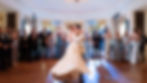 Bride and groom share a joyous dance as guests in elegant attire applaud. Chandelier above; band in the background adds to the festive mood.